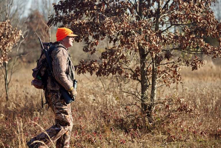 A hunter wearing a blaze orange hat walks in a field, consider bolt-action shotgun concept. 