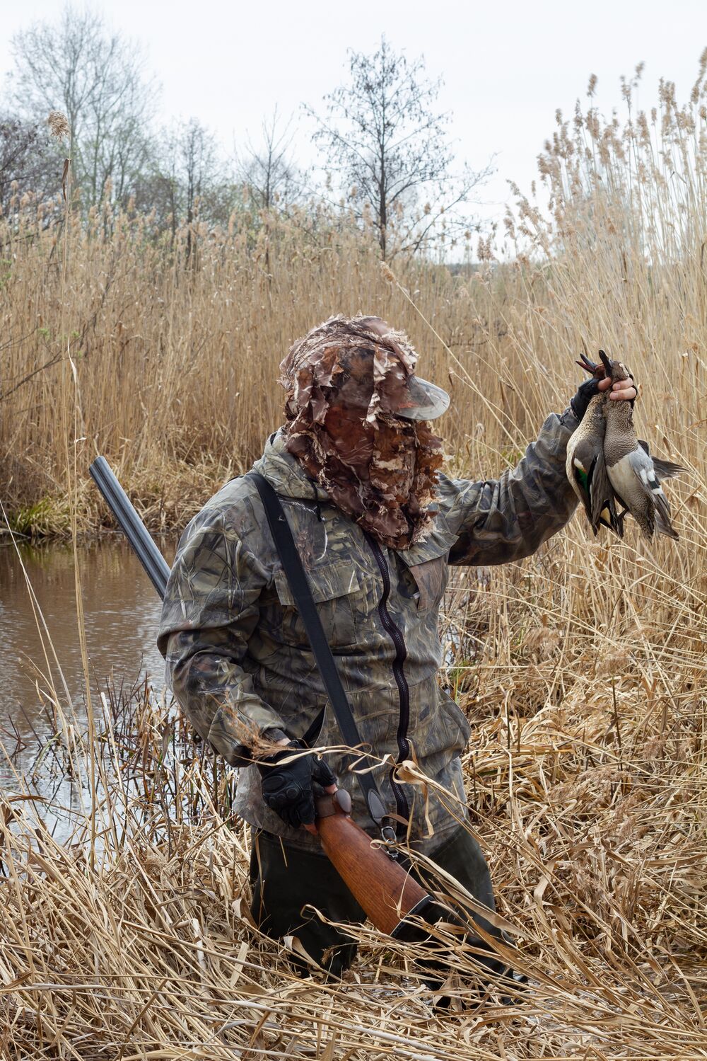 A hunter in camo at the edge of water holding a firearm and birds, understanding TABK concept. 