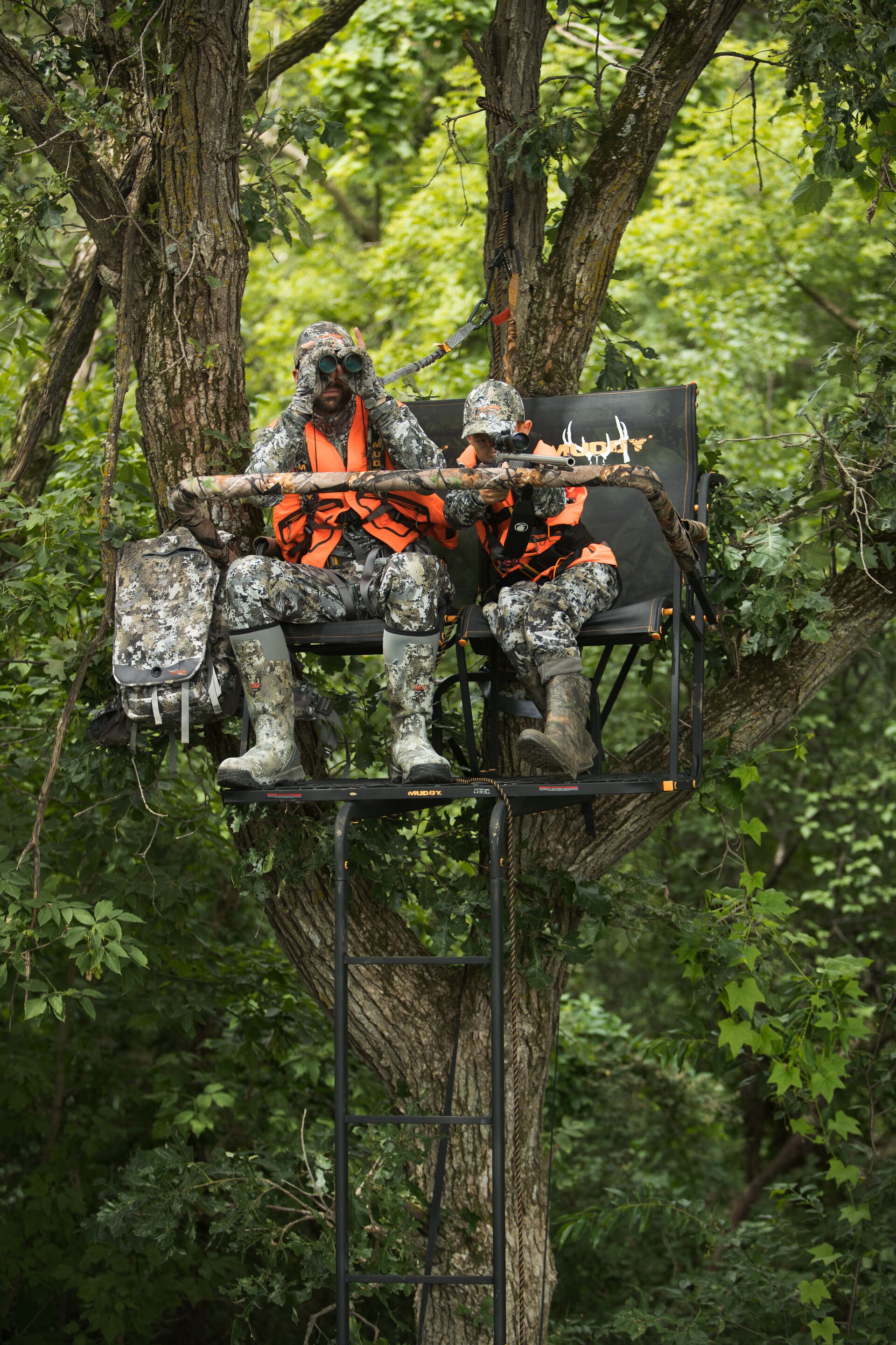  Man and young hunter in camo and blaze orange in a tree stand. 