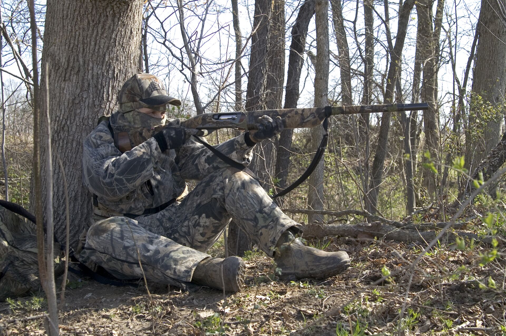 A hunter in camo against a tree aims a firearm, how to learn to hunt concept. 